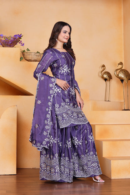 Woman in a purple traditional outfit with white patterns standing in a decorative indoor setting.