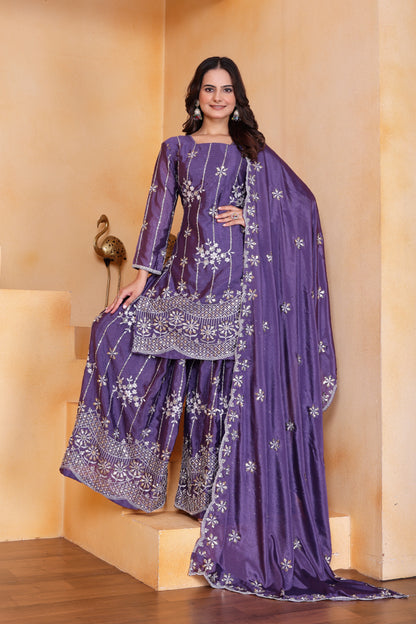 Woman in a purple traditional outfit with white patterns against a beige wall.