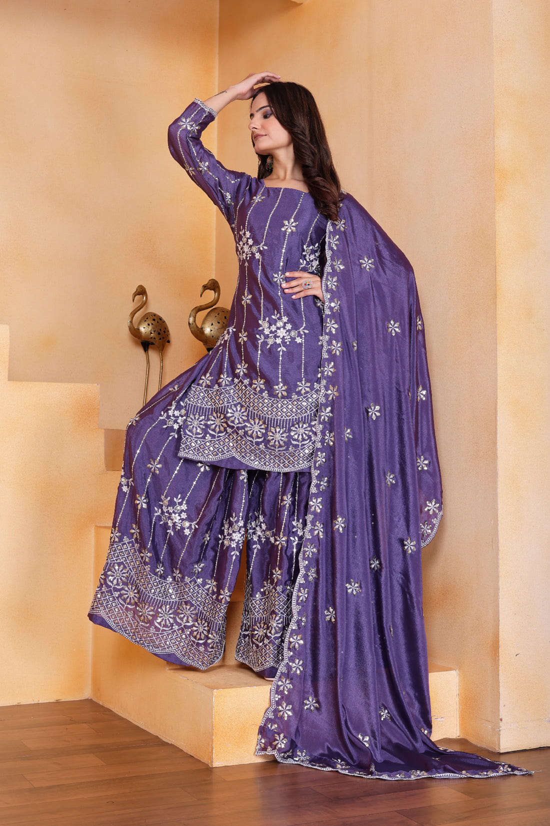 Woman in a purple traditional outfit with floral patterns against a beige wall.